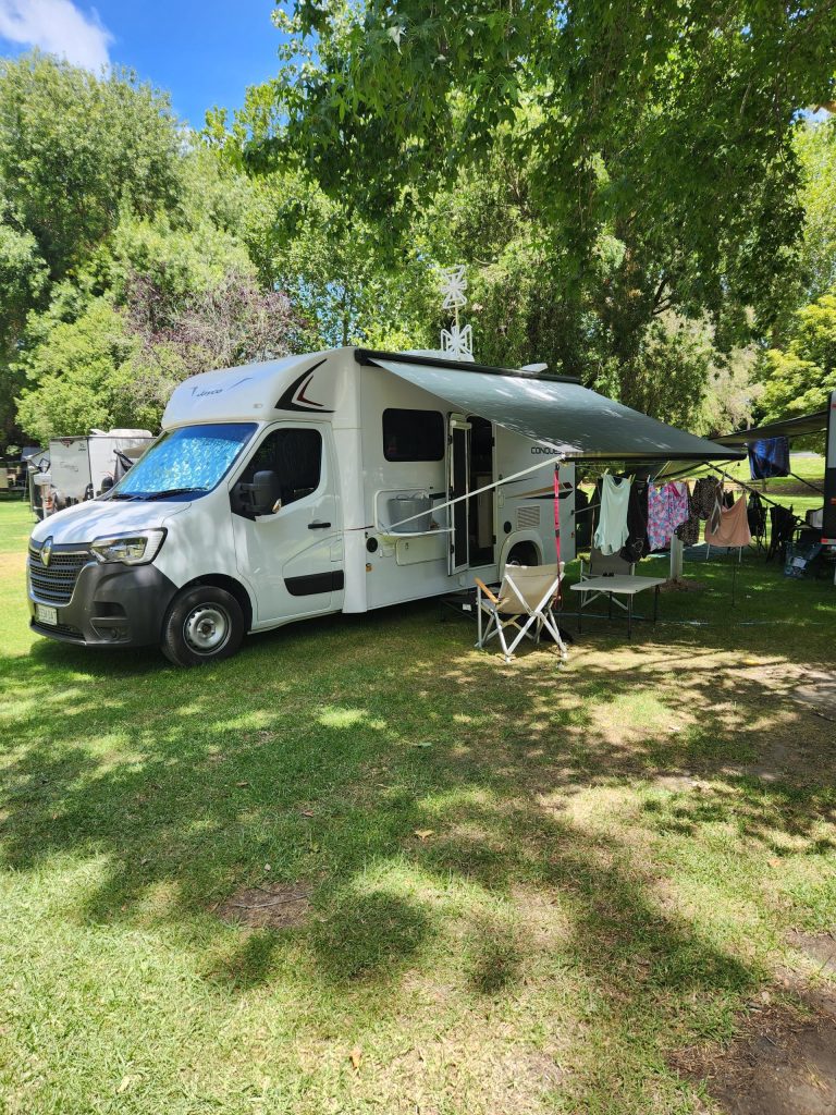 picture of a motor home parked on grass amongst green trees with a camping chair beside it under an awning